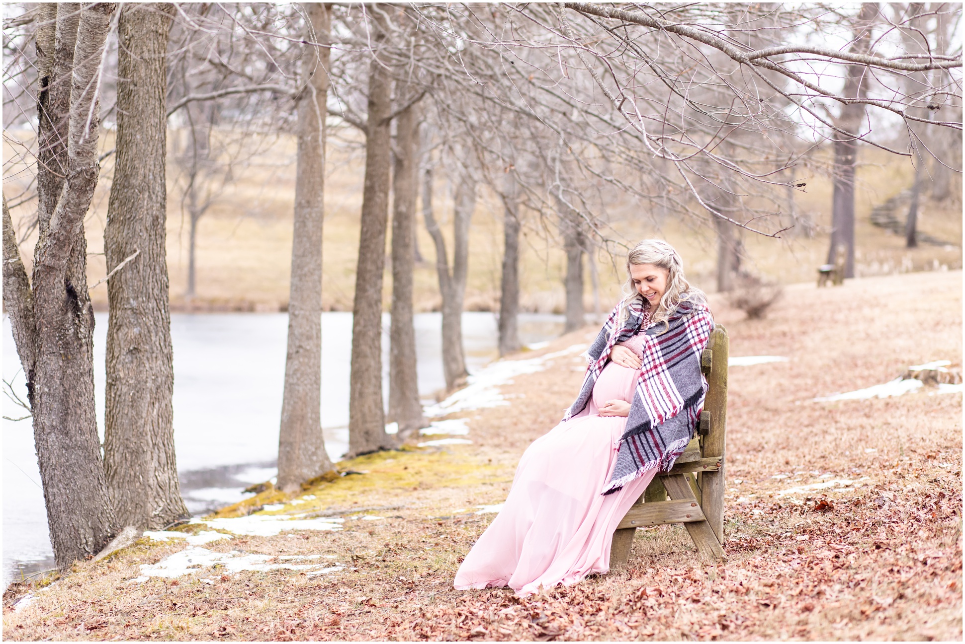 Horizontal Portrait of Pregnant Woman sitting on a bench by a pond holding her belly, wearing a winter shawl and a pink dress.