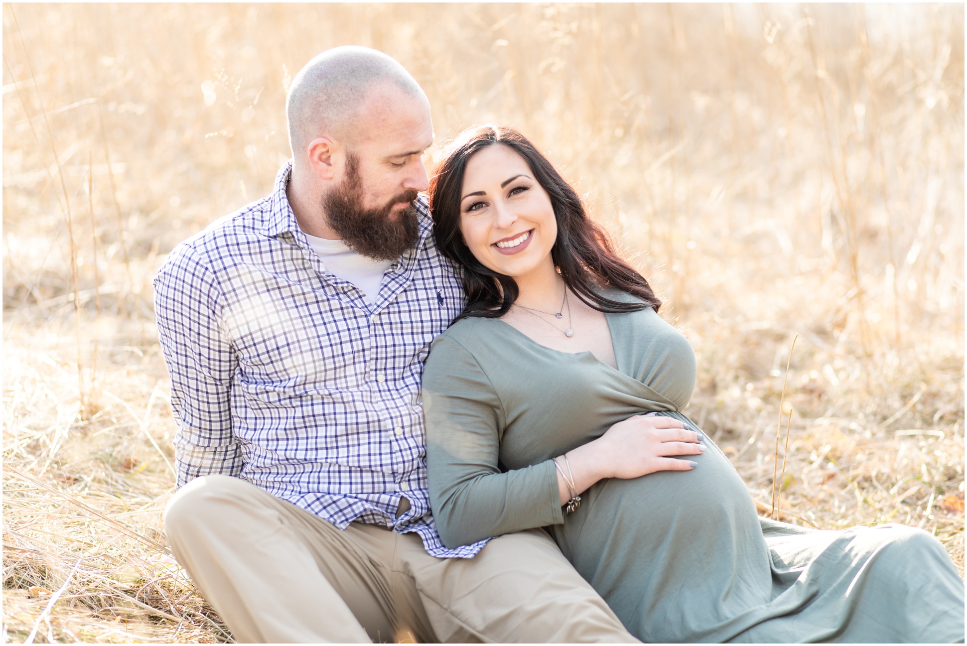 Husband and Wife sitting in the golden grass showing off their baby bump.