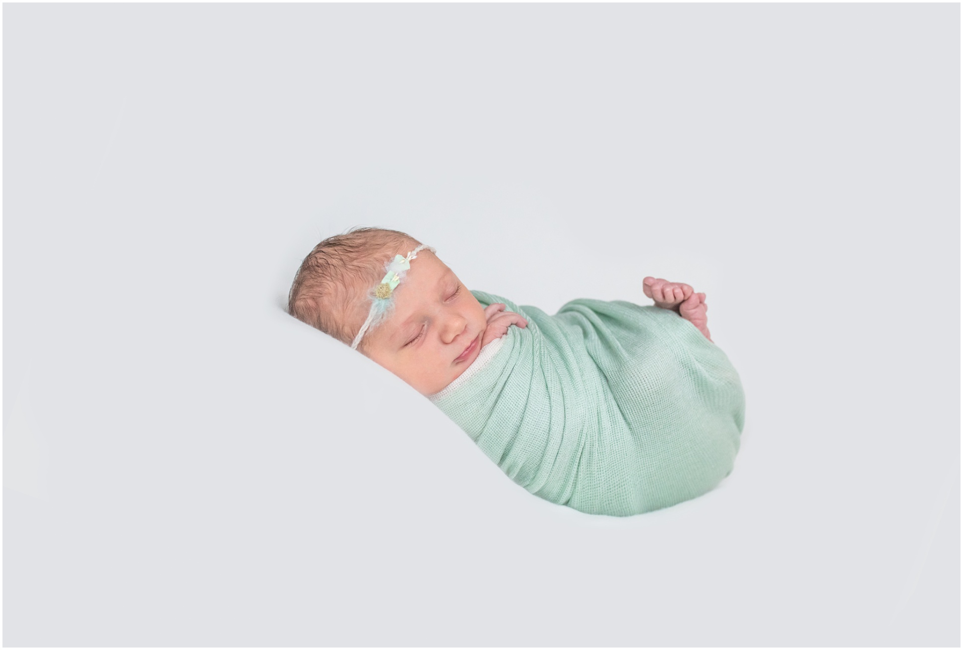Newborn curled up in a mint green wrap with a headbanc