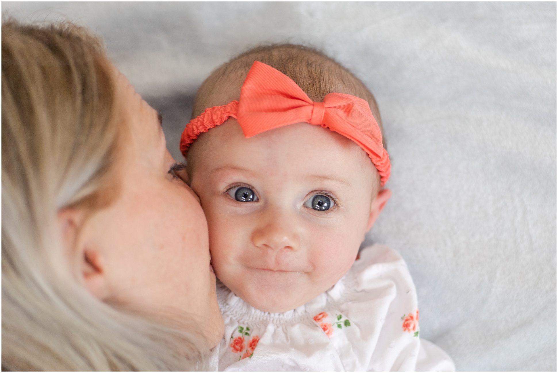 Lily Sage wearing a coral bow, smiling at the camera, while her mamma, Rebecca kisses her cheek