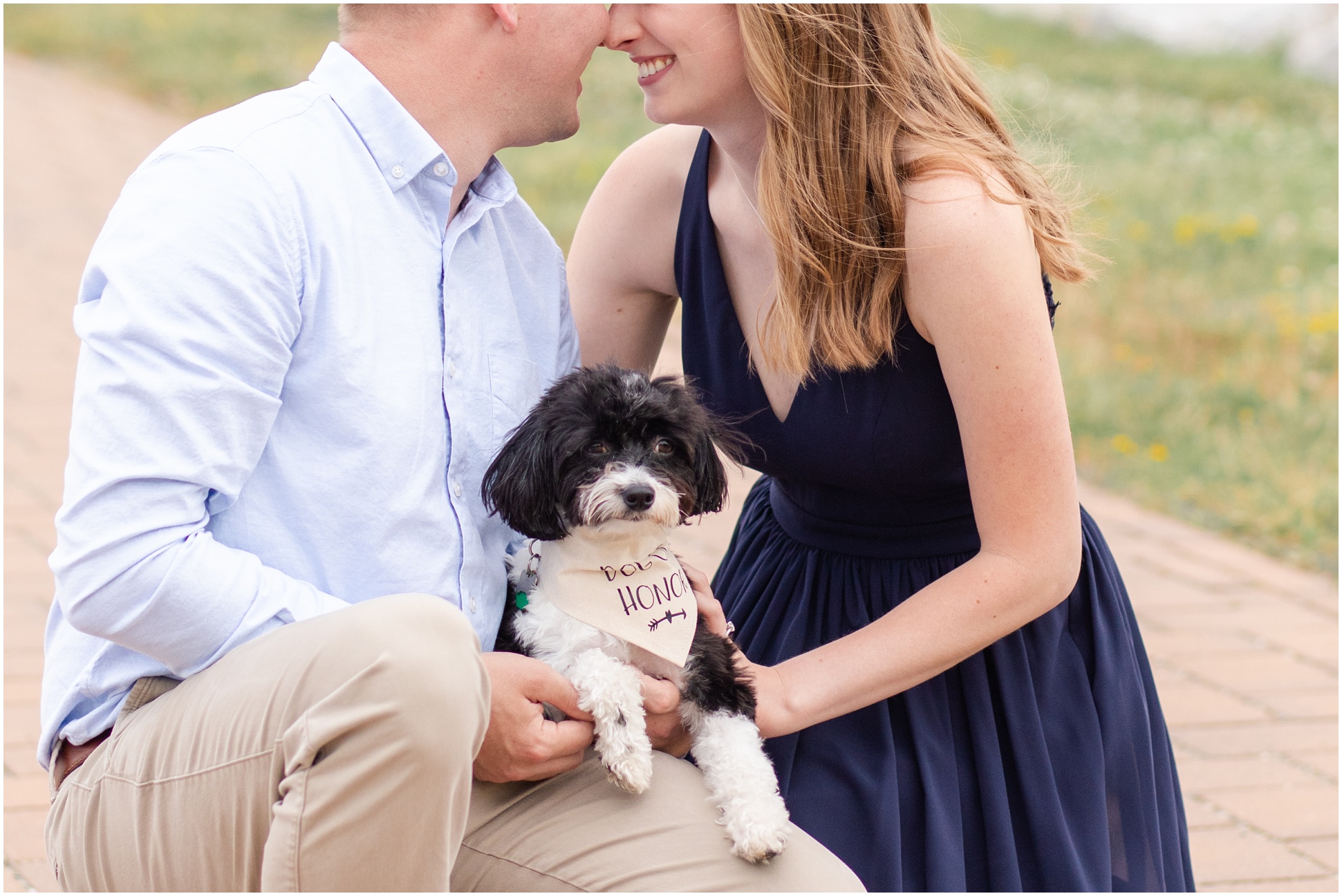 Black and White Dog sitting on engaged couples lap, while couple is nose to nose and puppy is wearing "Dog of Honor" bandana