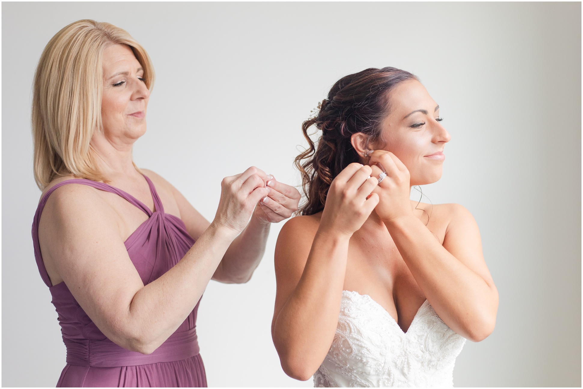 Mother of the bride wearing a purple dress, helping her daughter put on her wedding day earrings
