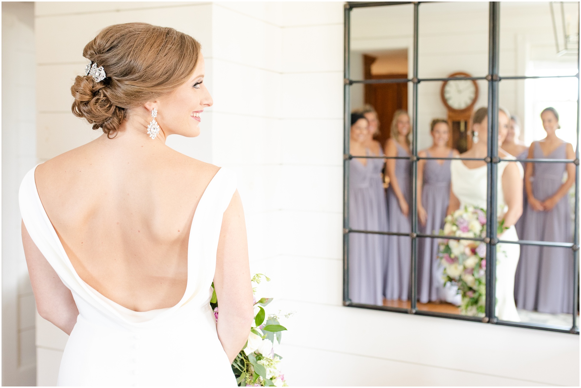 Bride wearing an open back dress, looking over her shoulder while her bridal party is looking at her through the mirror