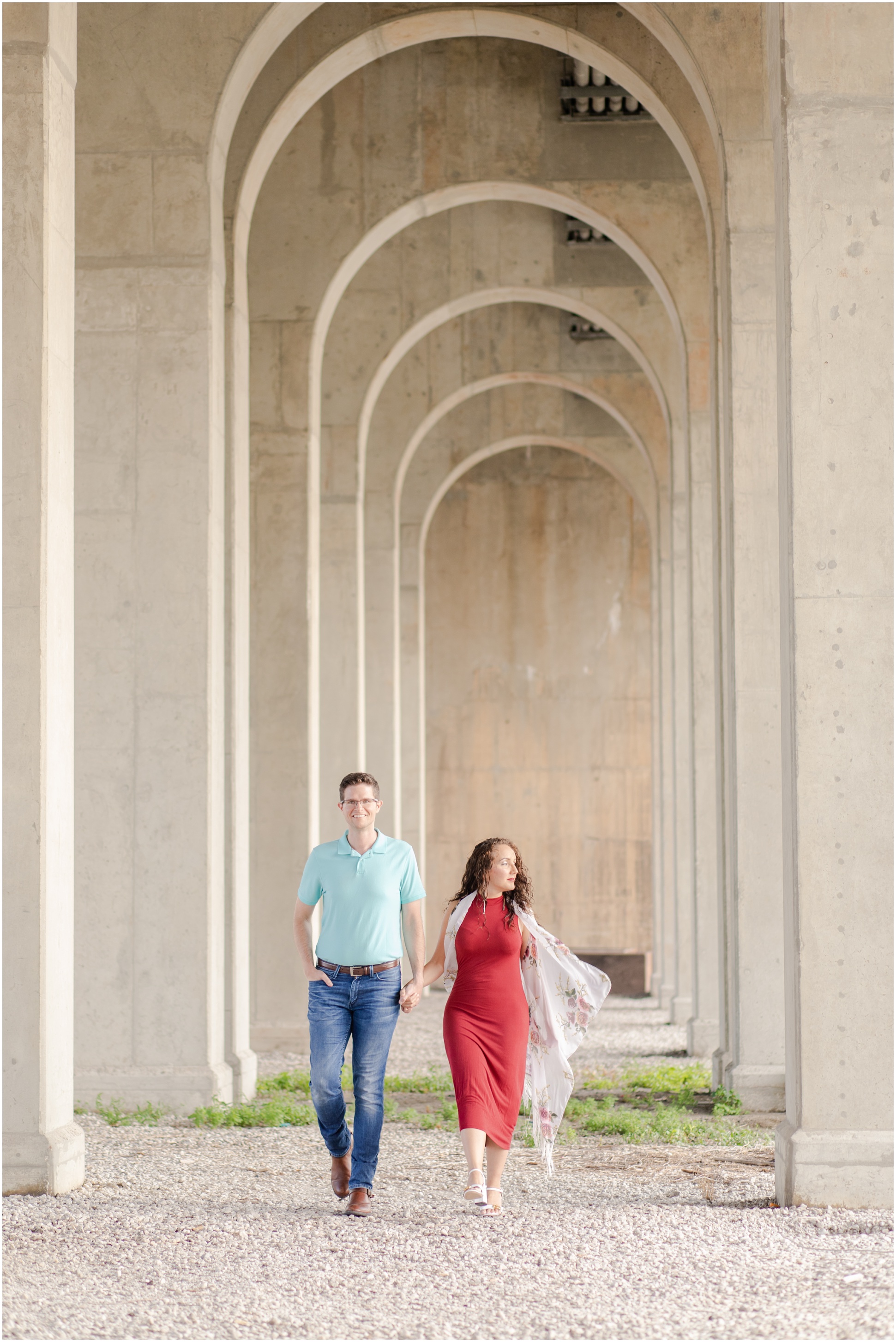 Man wearing teal shirt and woman wearing a red dress and a shaw walking hand-in-hand under The Hanover Street Bridge