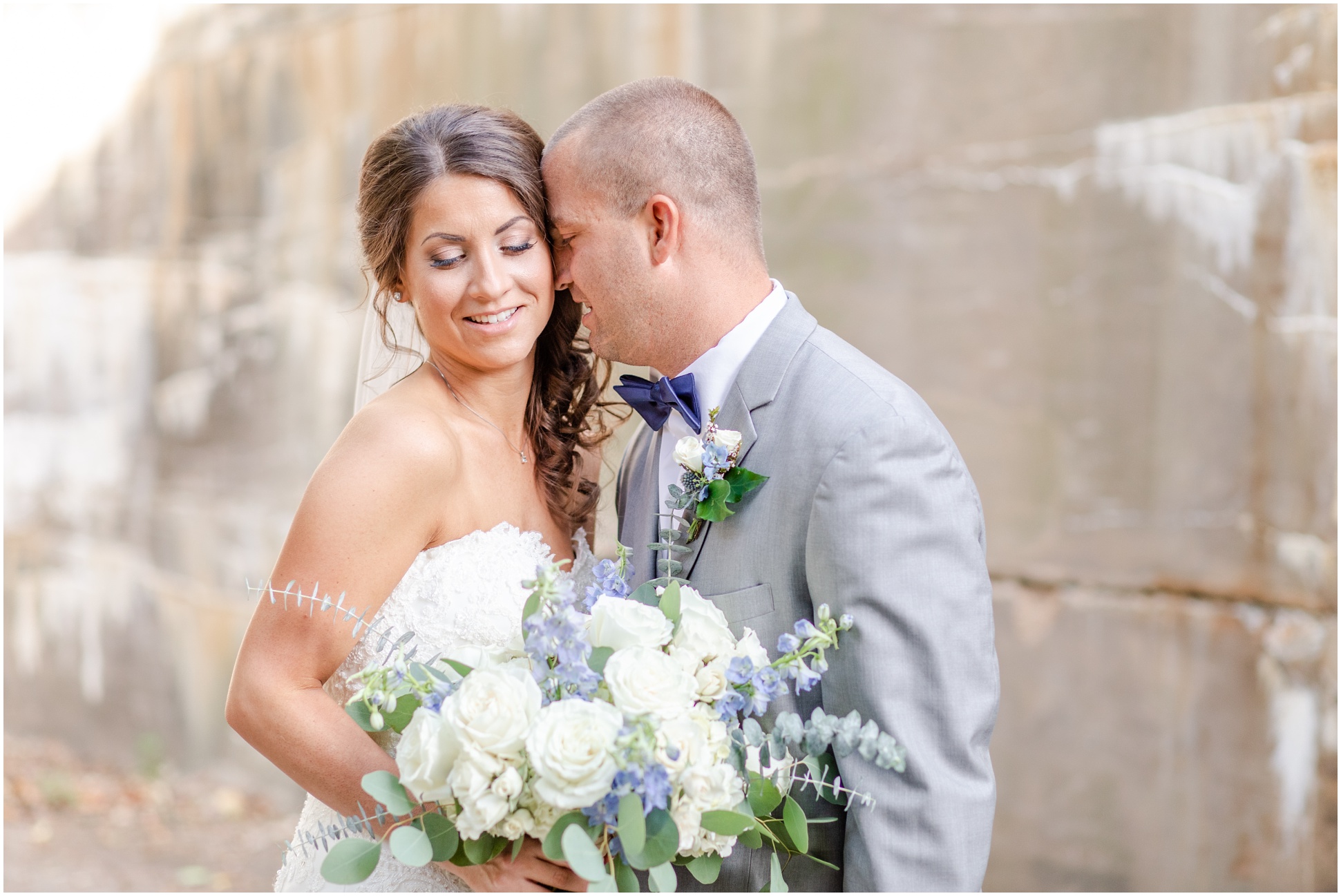 Groom nuzzling into the bride in front of a stone wall while holding her bouquet