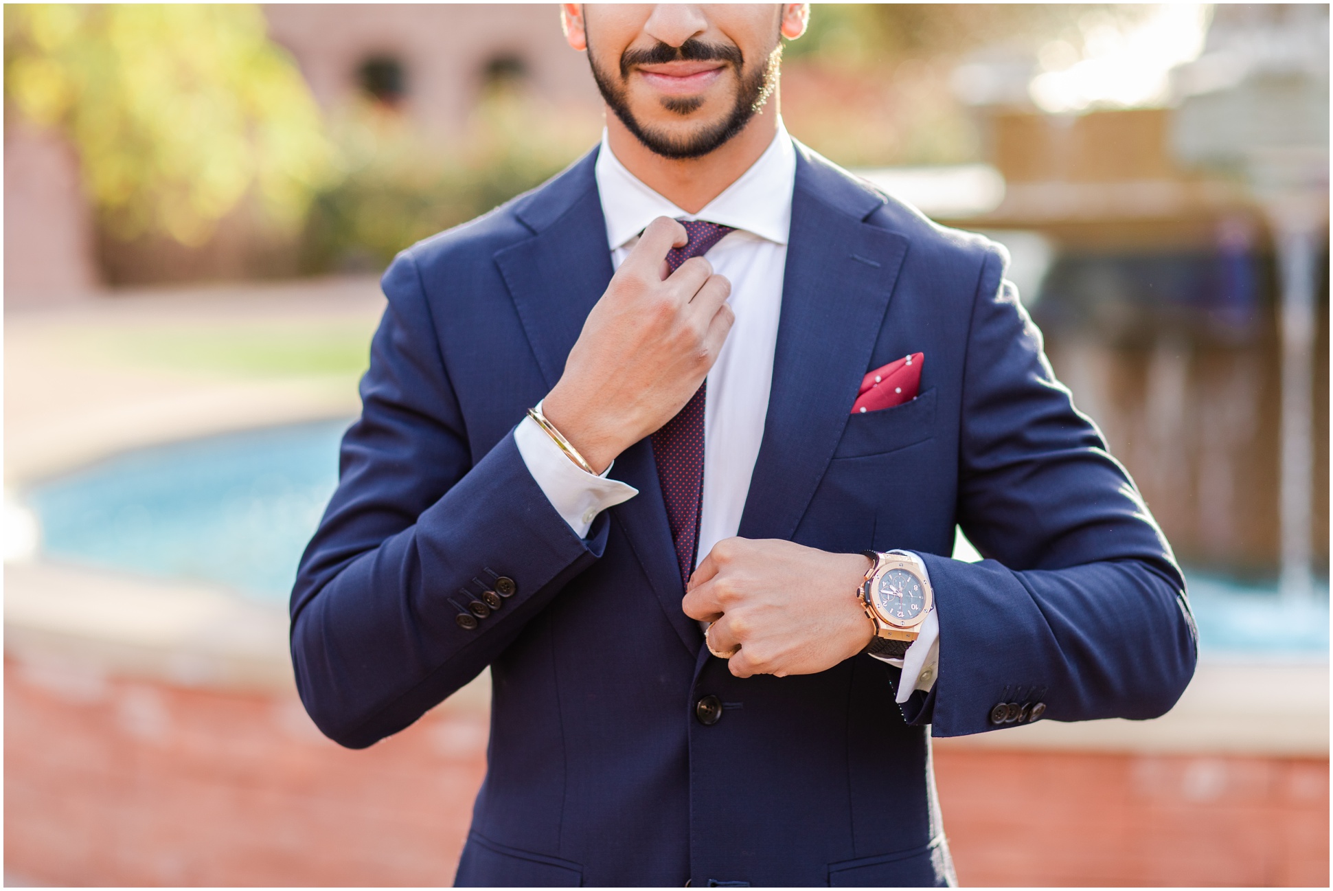 Young adult wearing navy blue suit with gold watch and fixing maroon tie