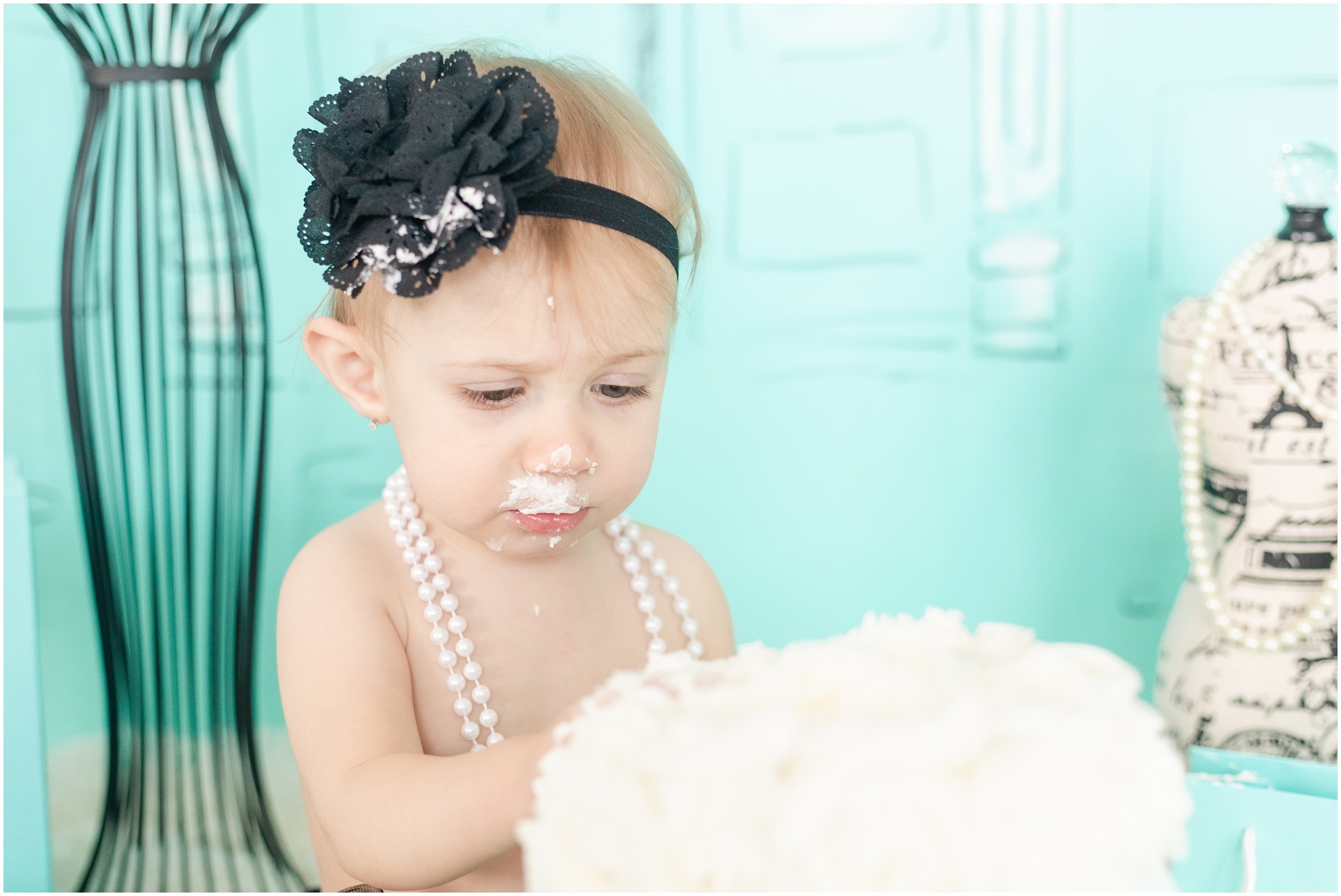 Lily in black flower headband and white pearls eating cake with frosting all on her face
