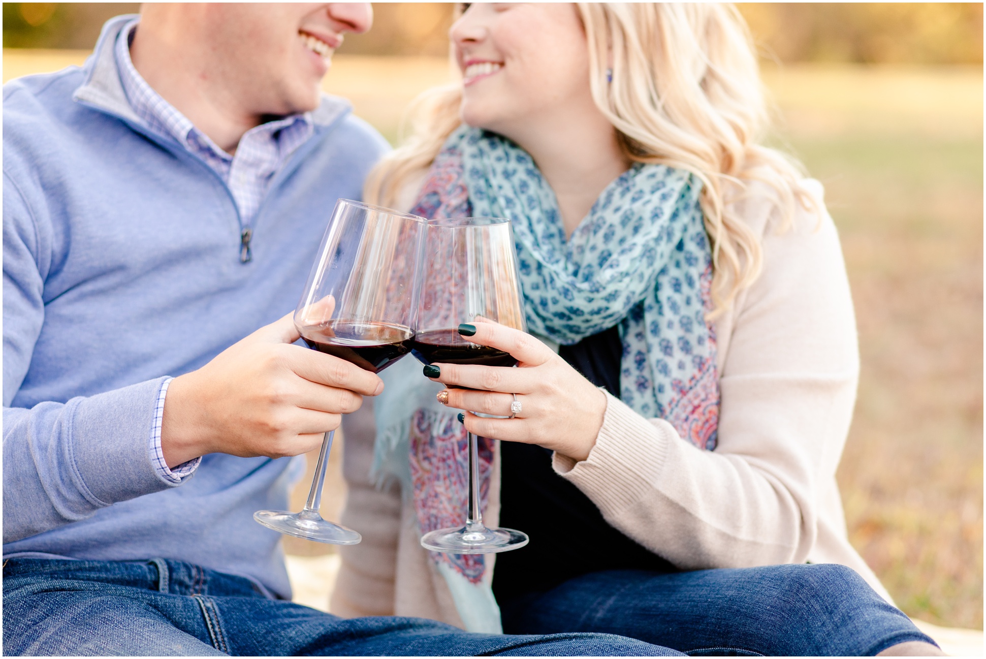 couple smiling while sitting on pick nick blanket while clicking wine glasses 