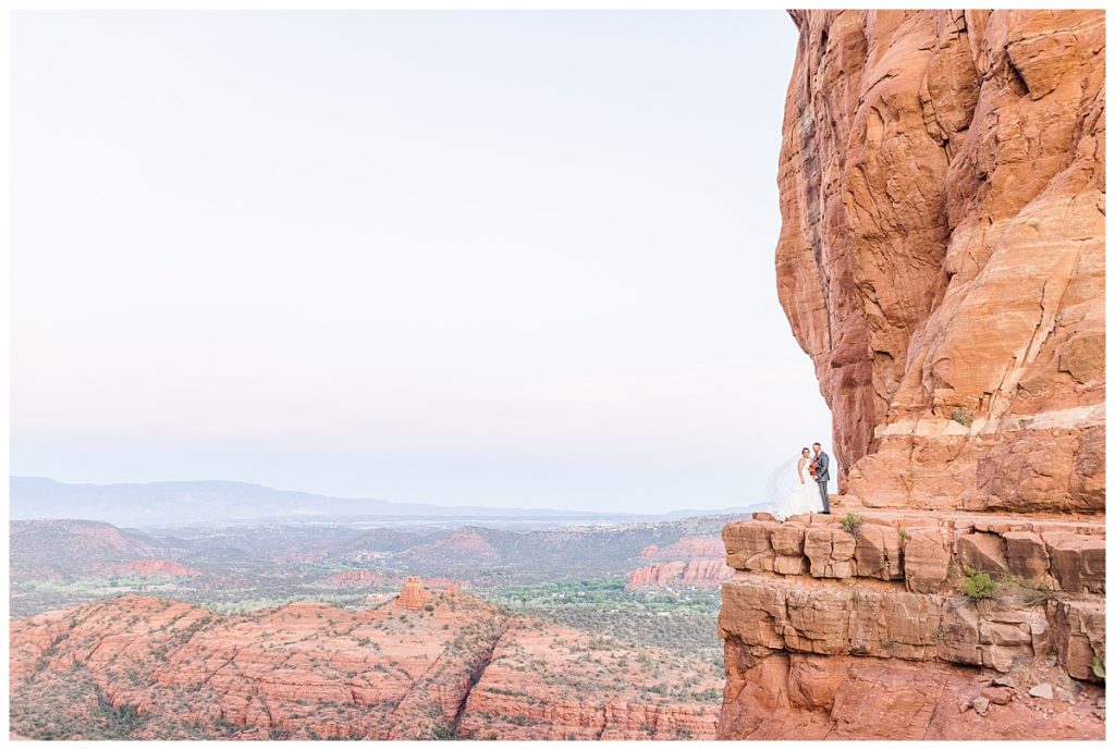 Epic wide shot of husband and wife in wedding attire standing on the ledge of Cathedral Rock in Sedona