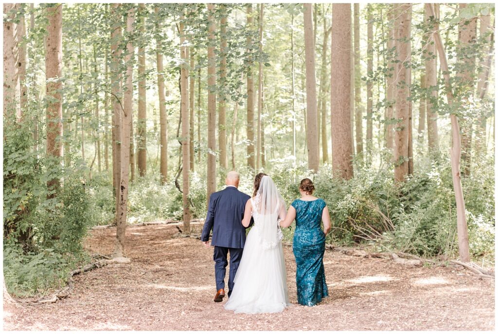 The bride's parents walk her down a forest path to the wedding ceremony